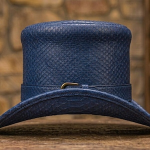 May include: A deep blue top hat featuring a textured snakeskin-like pattern across the entire surface. The hat includes a matching blue band wrapped around the base, secured with a small, metallic, brass-colored buckle at the front center.