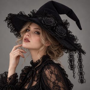 Può includere: Un cappello da strega nero caratterizzato da una tesa larga decorata con un intricato bordo in pizzo nero e due grandi rose in tessuto nero. Il cappello presenta una corona a punta e lunghi nastri di pizzo che pendono dal lato della tesa.