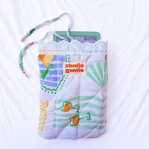 May include: A quilted fabric sleeve featuring a white background with a colourful print of lemons, green striped umbrellas, and wine glasses. The sleeve has a white eyelet lace trim at the top, fabric ties, and a label reading studio gwnio.