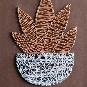 May include: A string art piece on a brown wooden board featuring a potted plant design. The plant leaves are created with orange thread wrapped around silver nails, while the semi-circular pot is formed with white thread and silver nails.