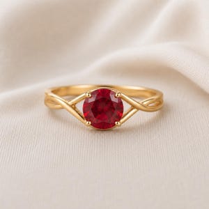 Puede incluir: Un anillo de oro con una piedra preciosa roja oscura, redonda y facetada, sujeta por un engaste de cuatro garras. La banda presenta un diseño dividido y trenzado que converge en la base de la piedra central, creando un aspecto simétrico.