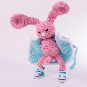 May include: A pink crocheted rabbit doll with long ears and black bead eyes. The rabbit wears a light blue crocheted cape with a white border and a pair of miniature light blue and white sneakers. The doll has a small tail and a pink nose.
