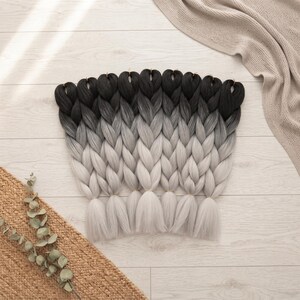 May include: A set of synthetic hair extensions featuring a distinct ombre colour transition. The strands start with a deep black at the top, blending into a medium grey in the middle, and finishing with a light silver-white at the bottom ends.