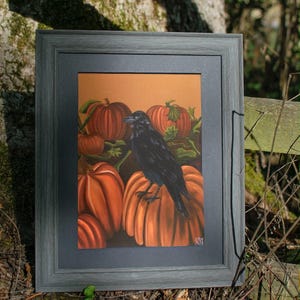 Può includere: Un dipinto incorniciato raffigurante un corvo nero appollaiato su una grande zucca arancione, circondato da altre zucche con steli e foglie verdi su uno sfondo arancione a tinta unita. L'opera è racchiusa in una cornice di legno grigio scuro.