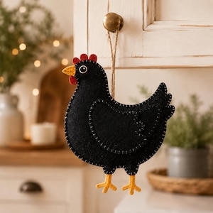 May include: A black felt chicken ornament featuring a yellow beak, red comb and wattle, and yellow feet. The chicken has white stitched detailing along the edges and is suspended by a thin brown twine loop attached to a round metal knob.