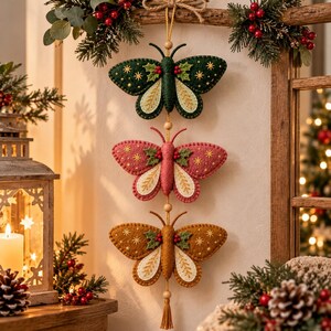 May include: A vertical hanging decoration featuring three felt butterflies in dark green, pink, and mustard yellow. Each butterfly is adorned with embroidered gold stars, green holly leaves, and red berries, connected by a string with wooden beads.