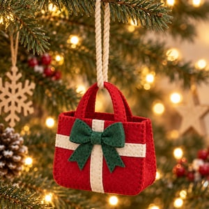 Pode incluir: Um enfeite de feltro vermelho em forma de pequena bolsa com duas alças e um laço de corda cor de creme para pendurar. A bolsa apresenta um design de listras verticais e horizontais de feltro creme e um laço de feltro verde escuro na frente.