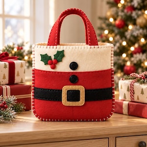 Pode incluir: Uma sacola de presente quadrada de feltro com design de traje de Papai Noel, apresentando uma base vermelha, um cinto preto com fivela bege, dois botões pretos e uma seção superior branca decorada com folhas de azevinho verdes e três bagas vermelhas.