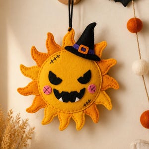 May include: A felt sun ornament featuring a yellow face with a black witch hat, jagged black eyes, a wide mouth with white teeth, and pink stitched cheeks. The sun has orange rays and black stitched details, hanging from a black cord loop.