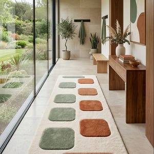 May include: A long rectangular runner rug with a cream-coloured base featuring a pattern of rounded square and rectangular shapes in muted sage green and burnt orange. The rug has a plush, textured pile and is positioned on a light stone floor.
