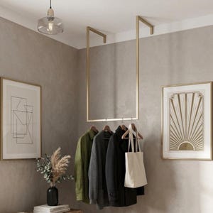 Pode incluir: Um cabideiro de metal dourado montado no teto com uma estrutura retangular. O cabideiro possui quatro cabides de madeira que sustentam uma camisa verde, um cardigã cinza, um casaco preto e uma bolsa de lona branca suspensos na barra.
