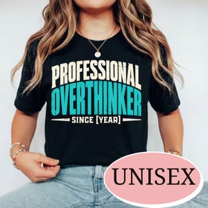 May include: A black short-sleeved t-shirt featuring the text PROFESSIONAL OVERTHINKER SINCE [YEAR] in a distressed font. The word PROFESSIONAL is in cream, while OVERTHINKER is in bright teal. A pink oval graphic with the word UNISEX is present.