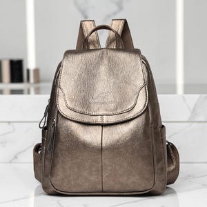Könnte beinhalten: Ein metallisch bronzefarbener Rucksack mit strukturierter Oberfläche, ausgestattet mit einer Klappe, einem Känguru-Logo und geprägtem Text, zwei Seitentaschen, einem Tragegriff sowie verstellbaren Schultergurten mit passenden Reißverschlüssen.