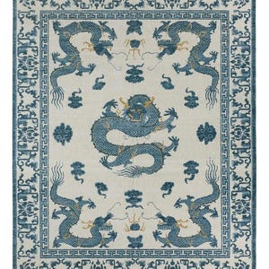 Op de afbeelding: Een rechthoekig vloerkleed met een crèmekleurige achtergrond, een centraal blauw drakenmotief en vier kleinere blauwe draken in de hoeken. De rand toont een geometrisch meanderpatroon en complexe blauwe bloem- en rankmotieven.