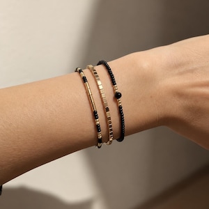 May include: A set of three bracelets featuring a combination of small black and gold-toned beads. One bracelet consists of square gold-toned beads, while the other two are composed of alternating black and gold-toned spherical beads.