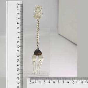 Può includere: Una forchetta in metallo color argento con manico intrecciato e una figura decorativa sulla sommità. Un righello in plastica trasparente è posizionato lateralmente e alla base, indicando che la forchetta misura circa 16,5 centimetri di lunghezza.