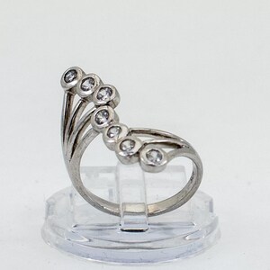 May include: A silver-toned ring featuring a curved, open-work design that rises to a point. The band is adorned with seven small, round, clear stones set in individual circular bezels, following the sweeping, asymmetrical arc of the metal frame.