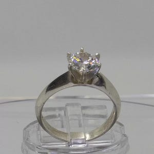 Op de afbeelding: Een zilverkleurige metalen ring met een enkele ronde, heldere, gefacetteerde edelsteen in een zetting met zes pootjes. De ringband is glad en loopt iets taps toe naar de zetting, met een reflecterend oppervlak en subtiele lichtreflecties.