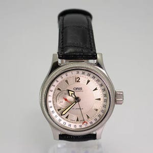 Puede incluir: Un reloj de pulsera automático Oris con esfera plateada, que presenta un anillo de fecha numerado del 1 al 31, una subesfera de segundos, manecillas luminosas y un centro plateado texturizado. Caja plateada pulida y correa de cuero negro.