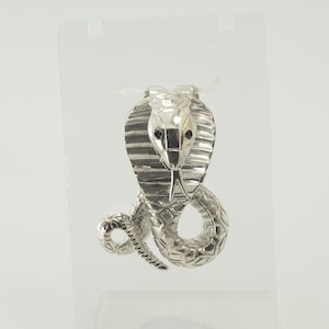 Op de afbeelding: Een zilverkleurige cobra-broche met een uitlopende kap met horizontale ribbels en een opgerold lichaam met een gestructureerd schubbenpatroon. De slang heeft twee kleine, donkere, ingezette ogen en een gespleten tong op een standaard.