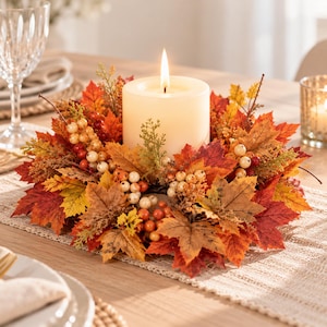 May include: A cylindrical cream-colored pillar candle sits in the center of a circular wreath made of artificial autumn foliage. The wreath features a mix of red, orange, and yellow maple leaves, small green sprigs, and clusters of white and red berries.