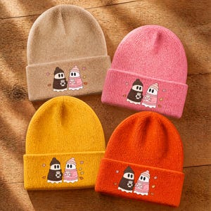 May include: Four knit beanies in tan, pink, yellow, and orange. Each hat features a graphic of two ghosts wearing winter hats and sweaters, holding mugs. The ghosts are positioned on the folded cuff of each beanie, surrounded by small autumn leaves.