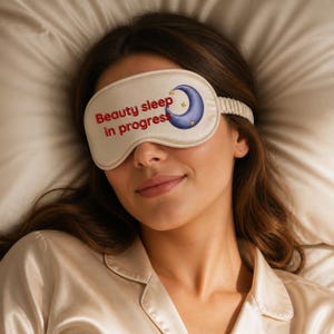 May include: A cream-coloured sleep mask featuring the red text "Beauty sleep in progress" and a graphic of a blue crescent moon with small yellow stars. The mask has an elasticated strap and is made of a smooth, satin-like fabric material.