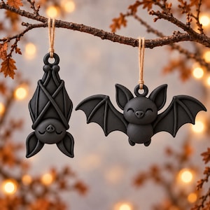 Könnte beinhalten: Zwei mattschwarze Fledermaus-Ornamente, die an einer braunen Paketschnur hängen. Eine Fledermaus ist hängend mit gefalteten Flügeln dargestellt, während die andere fliegend mit ausgebreiteten Flügeln zu sehen ist. Beide haben glatte Oberflächen.