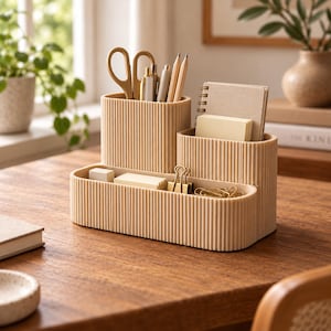May include: A beige desk organiser with a vertical ribbed texture, featuring multiple compartments for stationery. It holds a pair of scissors, pens, pencils, a small spiral-bound notebook, sticky notes, and several gold-toned metal binder clips.