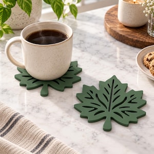 May include: Two dark green leaf-shaped coasters with intricate cut-out vein patterns. One coaster supports a speckled cream-colored ceramic mug filled with dark coffee, while the other sits empty on a white marble surface next to a striped cloth.