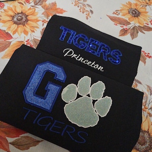 Puede incluir: Dos camisetas negras con diseños en azul y plata. La camiseta superior muestra la palabra TIGERS en lentejuelas azules sobre la palabra Princeton en letra blanca. La camiseta inferior presenta una gran letra G con purpurina azul, una huella plateada y la palabra TIGERS en azul.