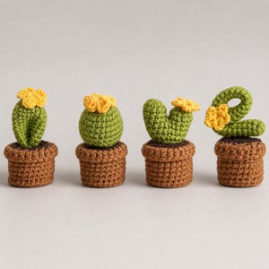 Op de afbeelding: Vier kleine gehaakte cactussen in bruine potjes. Elke groene cactus heeft een andere vorm en is bekroond met een klein geel bloemetje. De set bevat een hoge cactus, een ronde cactus, een cactus met twee takken en een gebogen cactus.