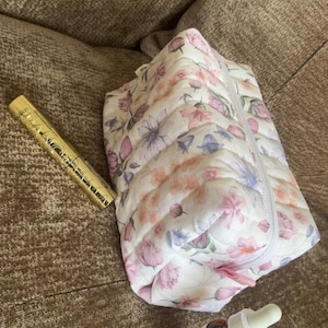 Peut inclure: Une trousse en tissu matelassé à fond blanc avec un imprimé floral doux dans des tons de rose, violet et vert. À côté se trouvent un tube de mascara cylindrique doré et un petit flacon compte-gouttes en verre brun étiqueté The Ordinary.