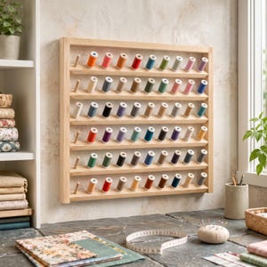 Op de afbeelding: Een rechthoekig, aan de muur gemonteerd garenrek van lichtgekleurd hout met zes horizontale planken. Elke plank bevat meerdere klossen naaigaren in diverse kleuren, waaronder rood, blauw, groen, geel, roze en verschillende neutrale tinten.