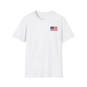 Könnte beinhalten: Ein schlichtes, weißes T-Shirt mit kurzen Ärmeln und einer kleinen, rechteckigen Grafik der amerikanischen Flagge im oberen linken Brustbereich. Das Flaggendesign hat eine leicht abgenutzte Pinselstrich-Optik in den Farben Rot, Weiß und Blau.