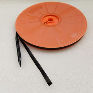 May include: A round orange plastic spool holding a long black garden tie. The black tie extends from the spool and features a pointed black plastic stake attached to the end, designed for securing plants or garden structures in an outdoor setting.