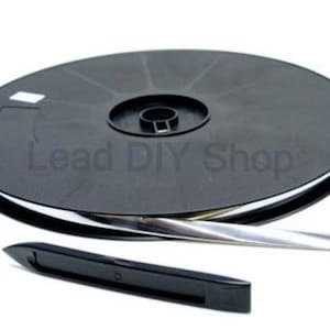 Könnte beinhalten: Eine schwarze Kunststoffspule mit einem langen, dünnen, metallisch silbernen Streifen, wobei der Text Lead DIY Shop auf der Spulenoberfläche sichtbar ist. Ein separates, längliches schwarzes Kunststoffwerkzeug mit spitzem Ende liegt davor.
