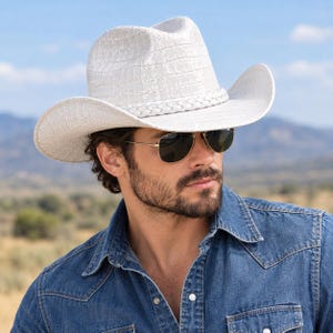 Op de afbeelding: Een witte cowboyhoed met een gestructureerd krokodillenleerpatroon en een gevlochten hoedband. De hoed heeft een brede, gebogen rand en een hoge kroon. Het wordt gedragen met een donkere pilotenbril en een blauw spijkerhemd met knopen.