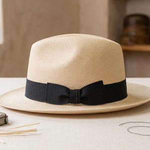 Può includere: Un cappello fedora in paglia intrecciata color beige chiaro con un'ampia fascia in nastro grosgrain nero e un fiocco centrale. Il cappello è posizionato su una superficie piana accanto a diverse bobine di filo in legno e un metro a nastro.