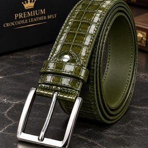Puede incluir: Un cinturón verde oliva brillante con textura de piel de cocodrilo y una hebilla de metal plateado. El cinturón cuenta con una trabilla de cuero a juego con un pequeño remache plateado. La caja es negra con el texto PREMIUM CROCODILE LEATHER BELT.