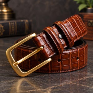 Pode incluir: Um cinto de couro castanho com textura brilhante de pele de crocodilo e uma fivela retangular de metal dourado mate. A tira do cinto tem uma série de orifícios de ajuste circulares e um passador de couro fixado por um rebite.