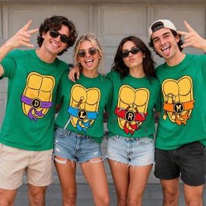 May include: Four green t-shirts featuring a yellow turtle shell graphic on the chest. Each shirt displays a unique coloured belt with a circular emblem containing the letters D, L, R, or M, and specific weapon illustrations like swords or nunchucks.