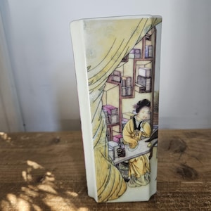 May include: A hexagonal ceramic vase featuring a detailed illustration of a person in yellow robes sitting at a desk with shelves of books in the background, framed by a draped yellow curtain design on a cream-coloured background.