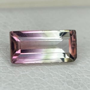 May include: A rectangular, baguette-cut gemstone featuring a distinct color gradient that transitions from a deep, saturated pink on one end to a clear, pale, and translucent tone on the opposite side, showcasing a faceted surface.