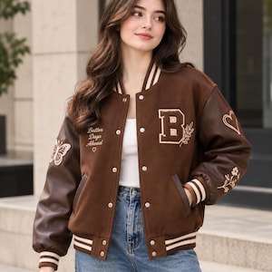 Puede incluir: Una chaqueta universitaria marrón con mangas de piel sintética marrón oscuro, con un parche de la letra B en blanco, diseños bordados de flores y mariposas, y el texto Better Days Ahead con un corazón. Tiene puños y dobladillo de canalé a rayas.