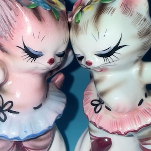 May include: Two ceramic cat figurines standing side by side. The left cat is pink with a blue tutu, and the right cat is white with brown stripes and a pink tutu. Both cats have painted closed eyes, long eyelashes, and floral headpieces.