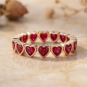 Könnte beinhalten: Ein Ewigkeitsring aus Roségold mit einer durchgehenden Reihe leuchtend roter, herzförmiger Edelsteine. Der Ring ist mit einer polierten Metallfassung gefertigt, die jeden einzelnen herzförmigen Stein sicher hält und ein kreisförmiges Design bildet.