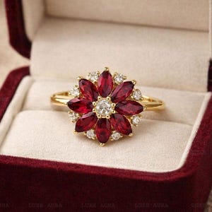 Könnte beinhalten: Ein Goldring mit einem blumenförmigen Cluster aus tiefroten Edelsteinen im Marquise-Schliff, die einen zentralen runden klaren Stein umgeben, mit kleinen runden klaren Steinen zwischen den Blütenblättern, in einer dunkelroten Samtschatulle.
