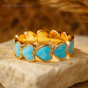 May include: A gold ring featuring a continuous band of connected heart-shaped light blue stones. Each heart is framed in a polished gold setting, creating a repeating pattern around the entire circumference of the jewelry piece.