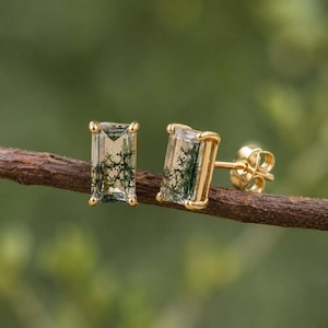 May include: A pair of rectangular stud earrings featuring moss agate gemstones with green dendritic inclusions, set in polished gold-toned metal prongs. Each earring has a clear, faceted stone surface and a standard post back closure mechanism.